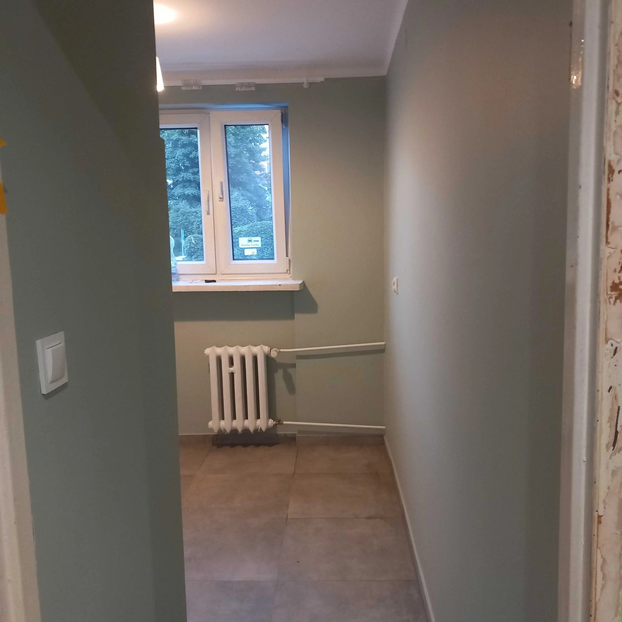 Korytarz po odświeżeniu, ściany pomalowane na oliwkowy odcień, widoczny grzejnik pod oknem i szare płytki podłogowe, futryna drzwi w trakcie renowacji.