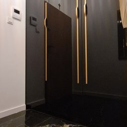 P.J. Patrycja Jaros - Czarna szafa wnękowa z wysokimi, złotymi uchwytami na tle błyszczącej, marmurkowej podłogi i białej ściany z panelem sterowania.