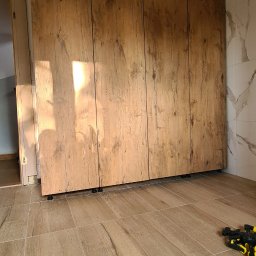 P.J. Patrycja Jaros - Montaż szafy wnękowej z drewnopodobnymi frontami, widoczne regulowane nóżki i fragment marmurowej ściany w tle.