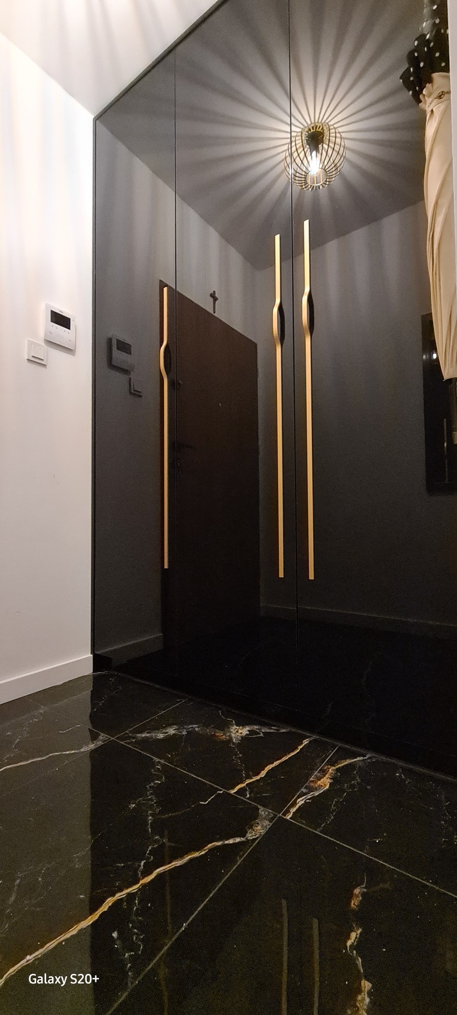 Czarna szafa wnękowa z wysokimi, złotymi uchwytami na tle błyszczącej, marmurkowej podłogi i białej ściany z panelem sterowania.