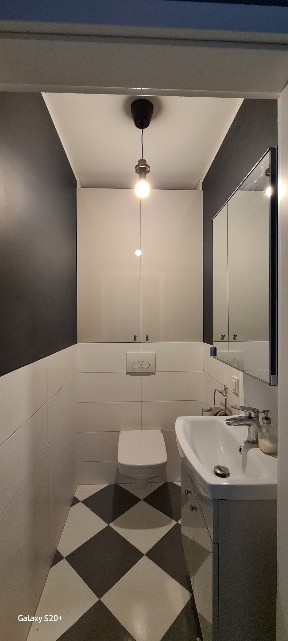 Wnętrze małej łazienki z białą ceramiką, zabudową nad toaletą, czarno-białą szachownicą na podłodze i szarą ścianą. Uwagę zwraca wisząca lampa z żarówką na długim kablu.