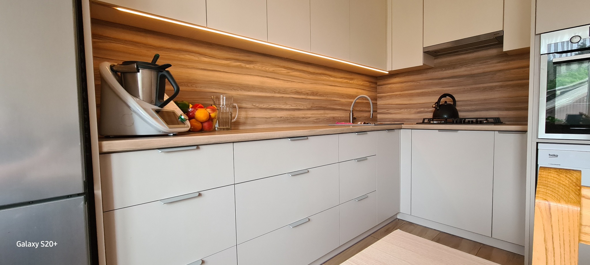 Nowoczesna, jasna kuchnia z drewnianym panelem ściennym, widoczne białe szafki z podłużnymi uchwytami, sprzęt AGD: Thermomix, czajnik, piekarnik Beko oraz owoce w szklanej misie.