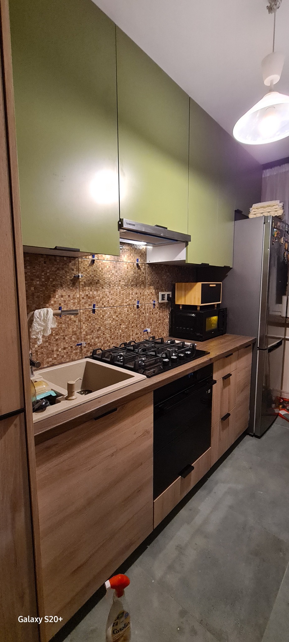 Fragment kuchni z zielonymi szafkami wiszącymi i drewnopodobnymi szafkami dolnymi, widoczna płyta gazowa, piekarnik, zlew z baterią, fragment lodówki i mozaiki na ścianie, z bliska widoczna butelka...