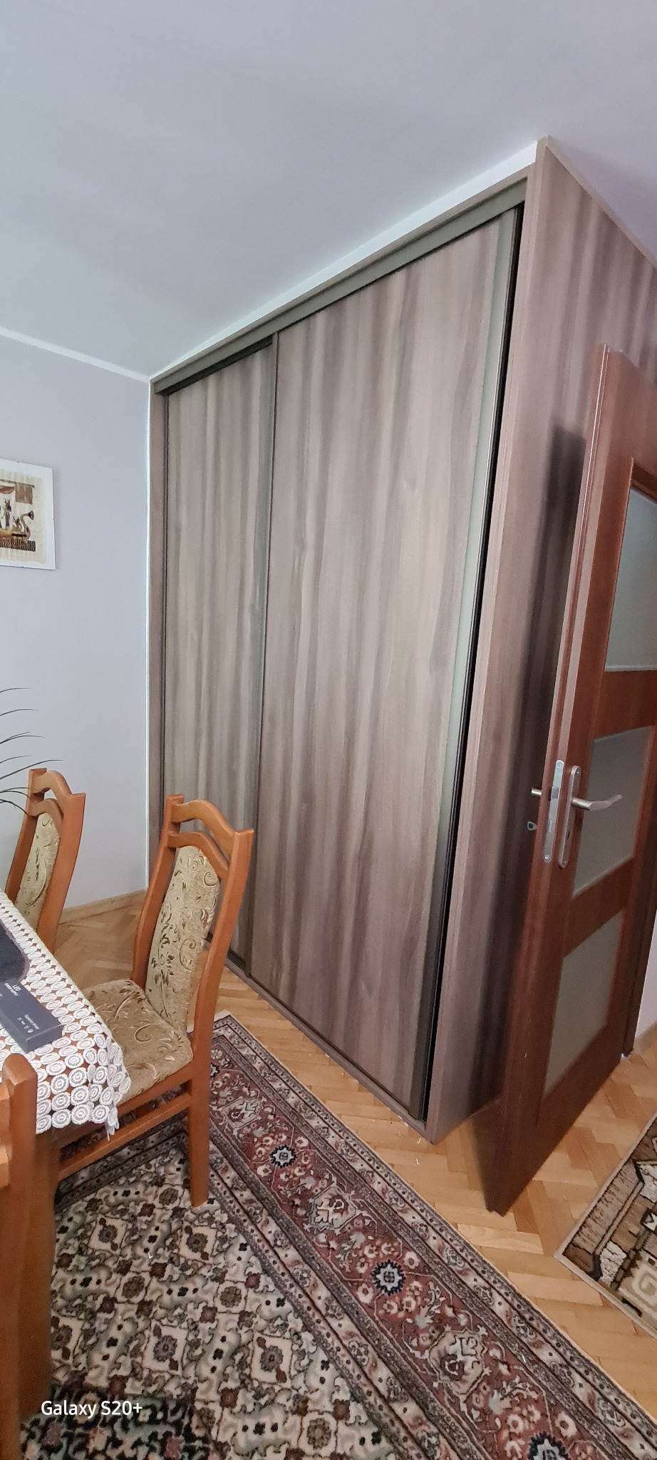 Wnętrze pokoju z dużą szafą przesuwną o drewnopodobnym wykończeniu, częściowo zasłoniętą otwartymi drzwiami, widoczny fragment stołu i krzesła na wzorzystym dywanie.
