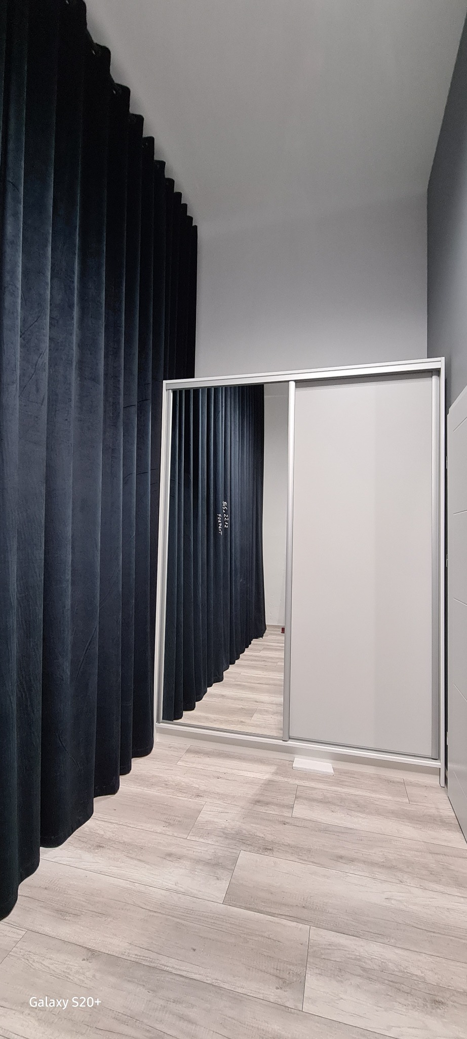 Wnętrze z szafą wnękową z przesuwnymi, lustrzanymi drzwiami, obok ciemne zasłony i panele podłogowe imitujące drewno.