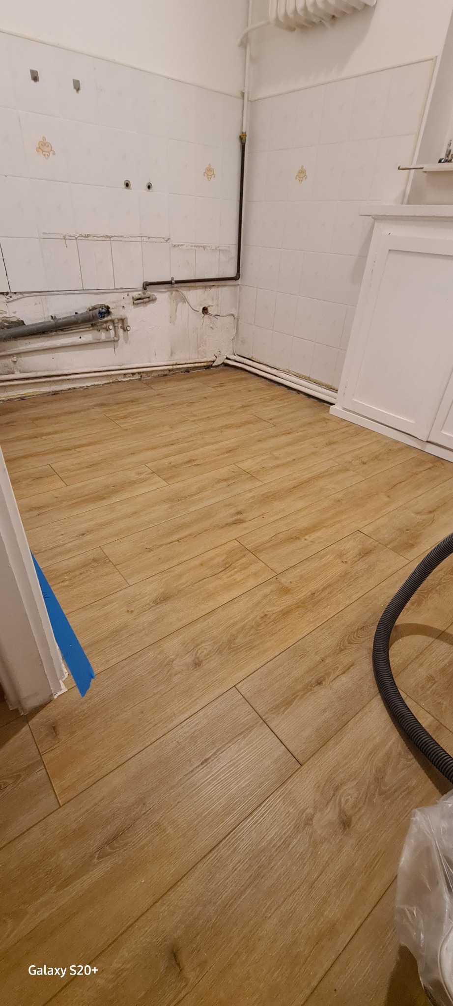 Beżowe panele podłogowe ułożone w kuchni przed montażem mebli, widoczne rury instalacyjne i fragment odkurzacza.