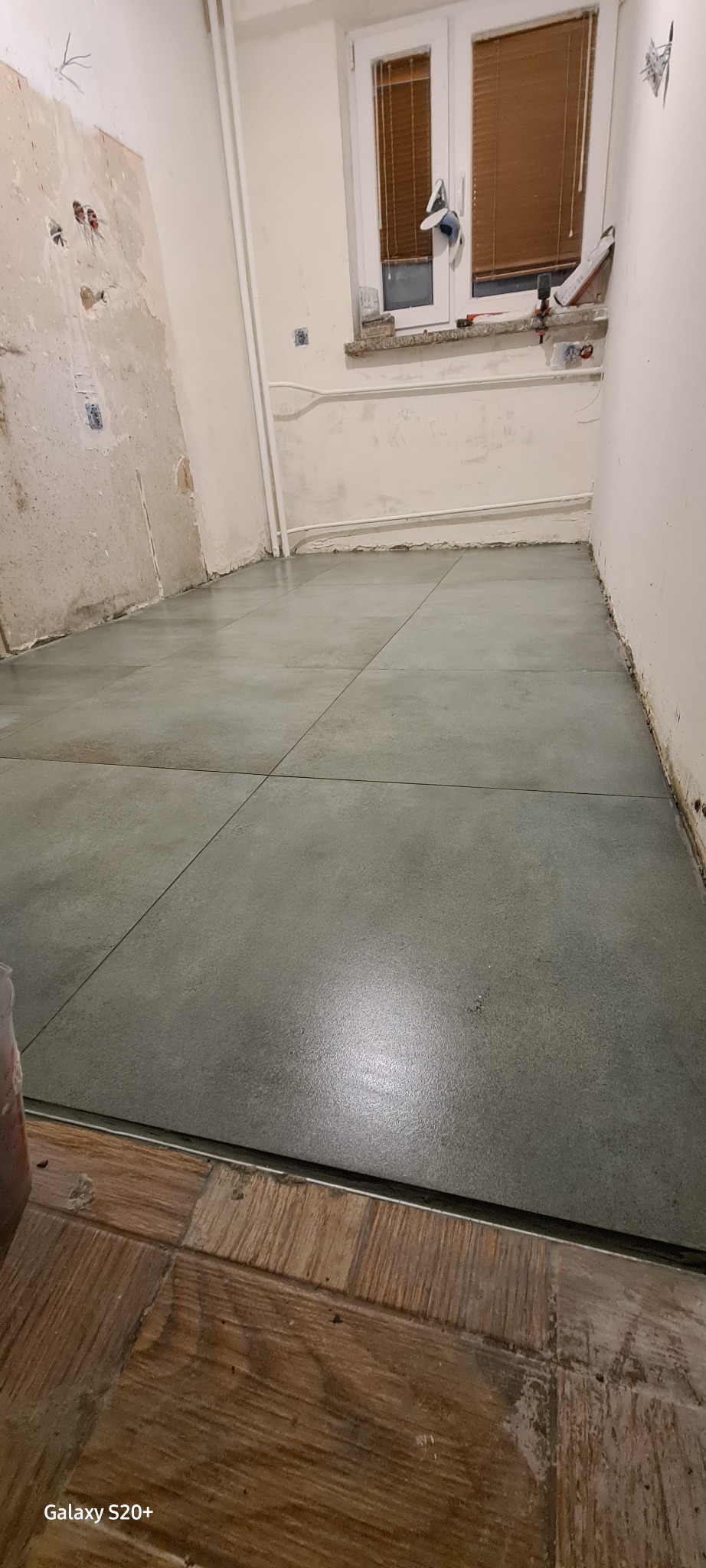 Szare panele podłogowe ułożone w pomieszczeniu w trakcie remontu, widoczne niedoskonałości ścian i instalacji elektrycznej, fragment progu z drewnopodobnych paneli.