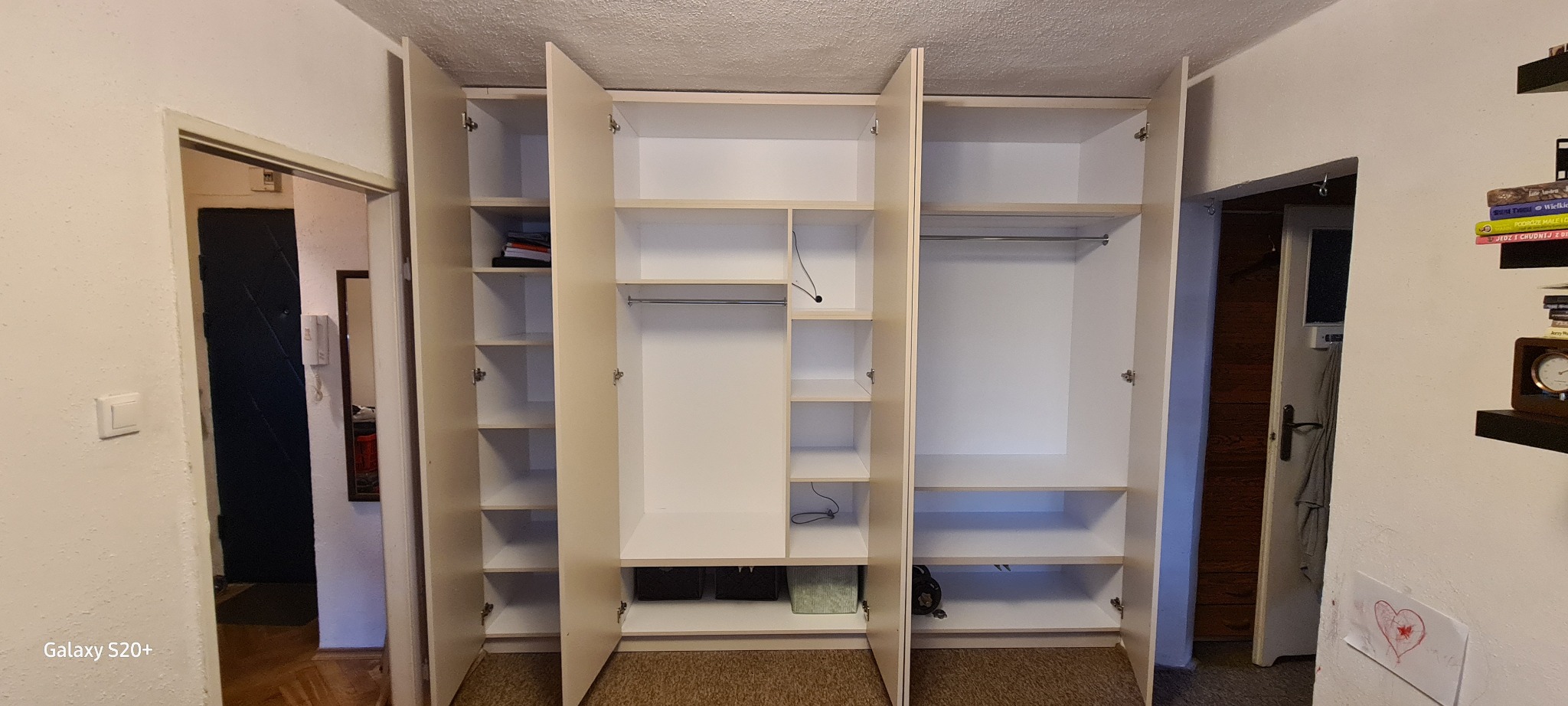 Wnętrze mieszkania z otwartą, trzydrzwiową szafą wnękową z jasnego drewna, z półkami po lewej stronie i drążkiem na ubrania po prawej, widoczne akcesoria w dolnej części oraz kable od oświetlenia.