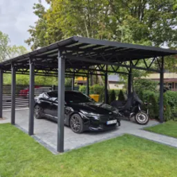 Nowoczesna, czarna konstrukcja zadaszenia nad miejscem parkingowym, z zaparkowanym czarnym samochodem i motocyklem pod pokrowcem, otoczona trawnikiem i kostką brukową.