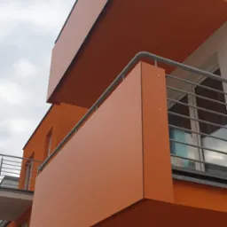 Nowoczesny budynek z pomarańczowymi balkonami z metalową balustradą, widok z dołu na tle pochmurnego nieba.