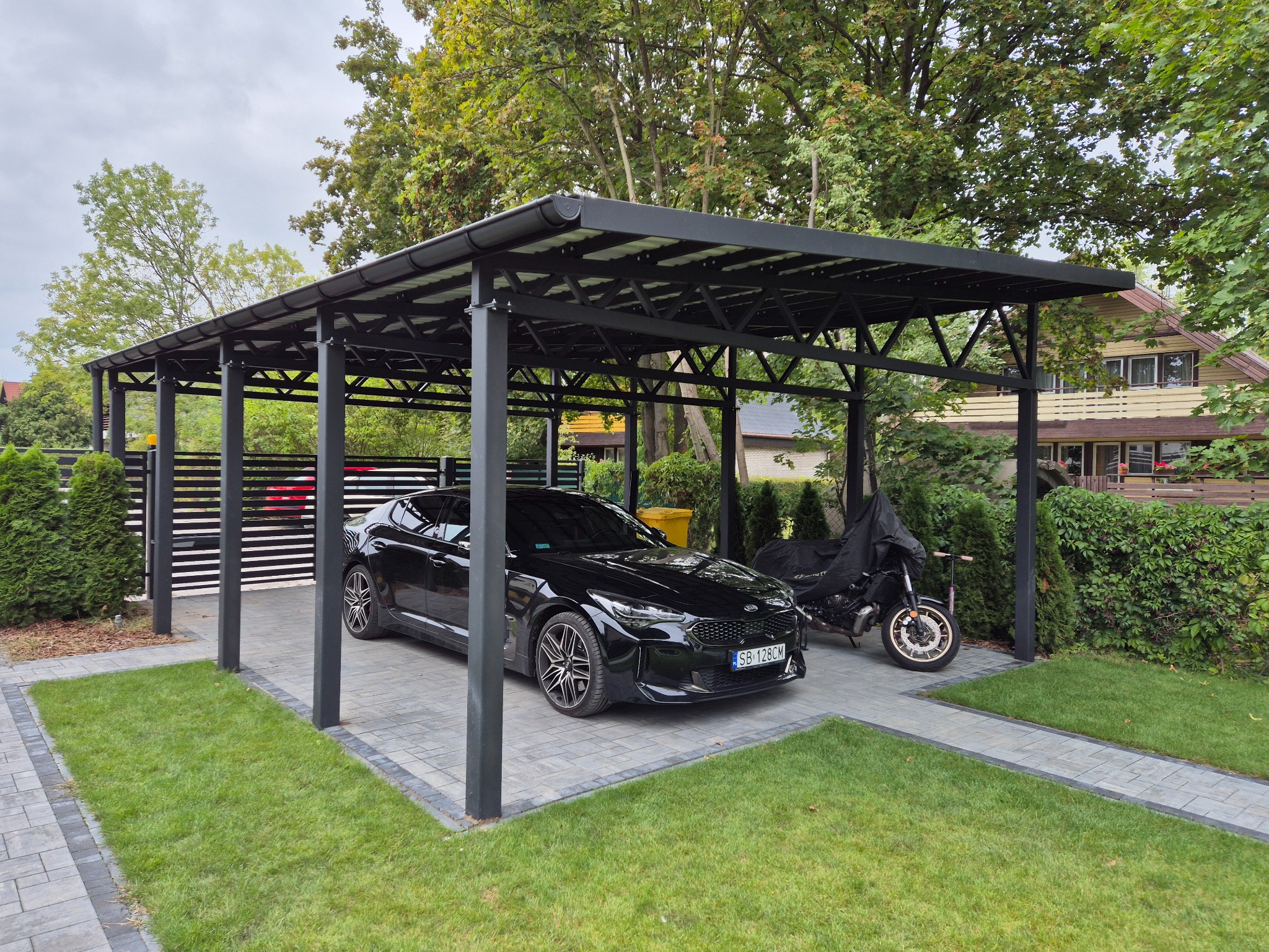 Nowoczesna, czarna konstrukcja zadaszenia nad miejscem parkingowym, z zaparkowanym czarnym samochodem i motocyklem pod pokrowcem, otoczona trawnikiem i kostką brukową.