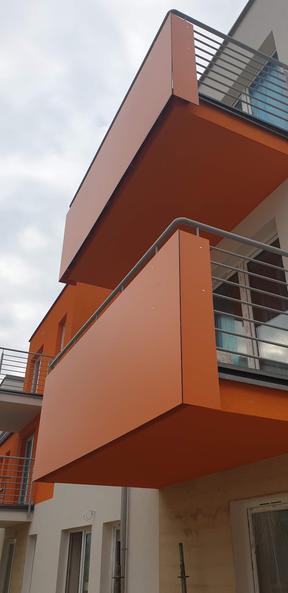 Nowoczesny budynek z pomarańczowymi balkonami z metalową balustradą, widok z dołu na tle pochmurnego nieba.