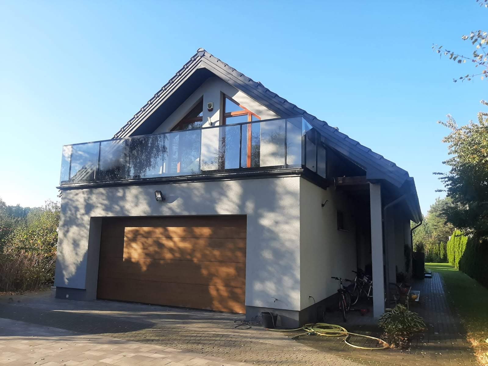 Nowoczesny dom z balkonem zabezpieczonym szklaną balustradą, drewniana brama garażowa, widok z zewnątrz w słoneczny dzień.