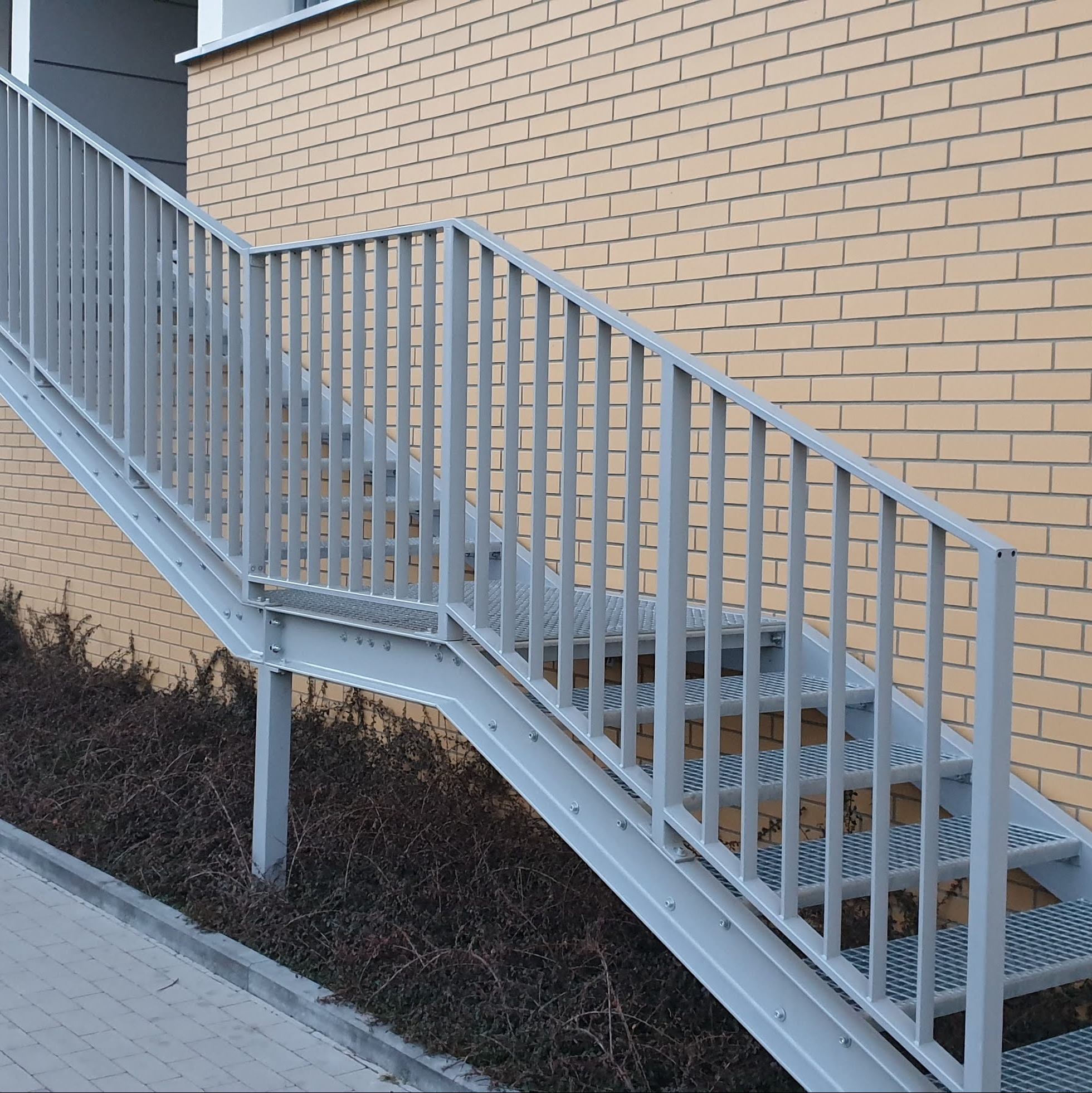 Zewnętrzne metalowe schody z ażurowymi stopniami i szarą balustradą, przylegające do ceglanego budynku.