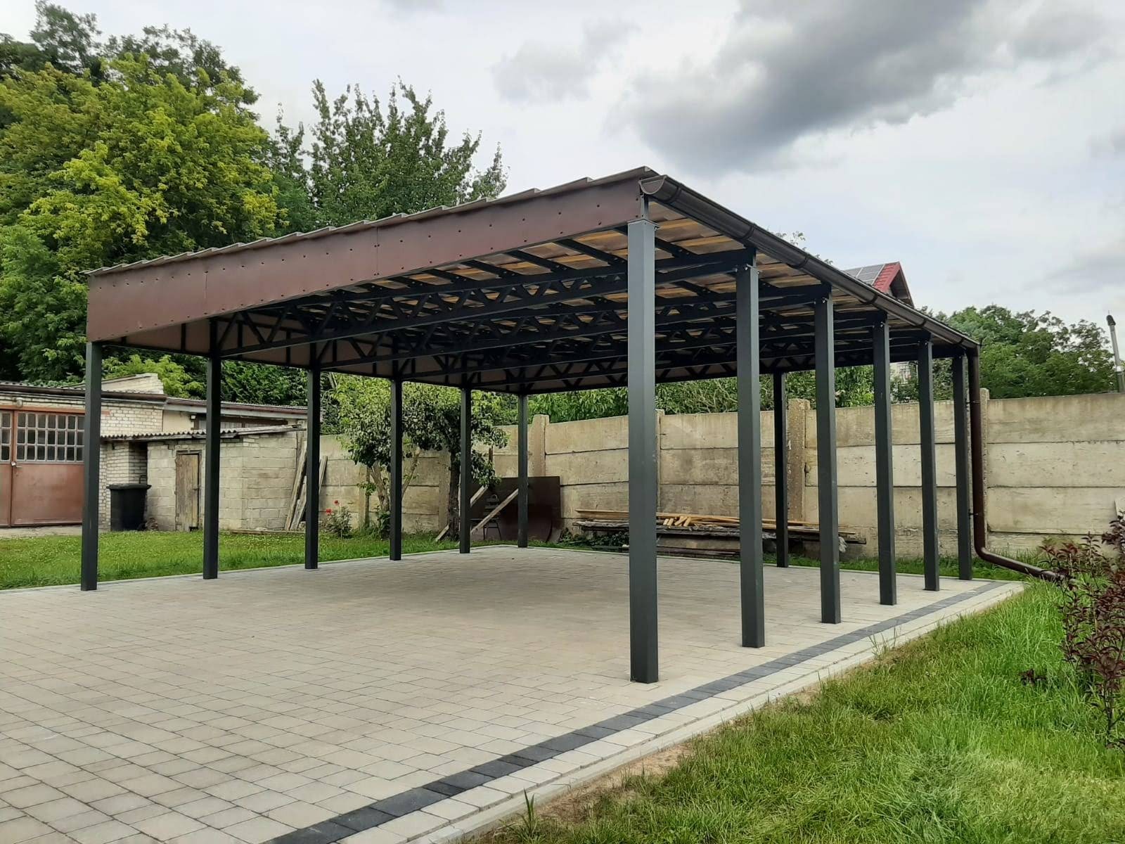 Zadaszona, metalowa pergola w kolorze brązowym z czarnymi słupami, na wybrukowanym podłożu, z widocznym fragmentem budynku gospodarczego w tle.