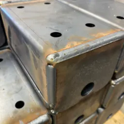 Stos metalowych, spawanych profili zamkniętych z otworami, widoczne ślady spawania i niewielka korozja powierzchniowa.