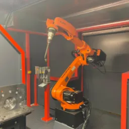 Pomarańczowy robot przemysłowy KUKA w szarej kabinie, z uchwytem narzędziowym skierowanym w stronę stanowiska pracy z metalowymi elementami, czerwona rama konstrukcyjna, stanowisko do obróbki metalu.