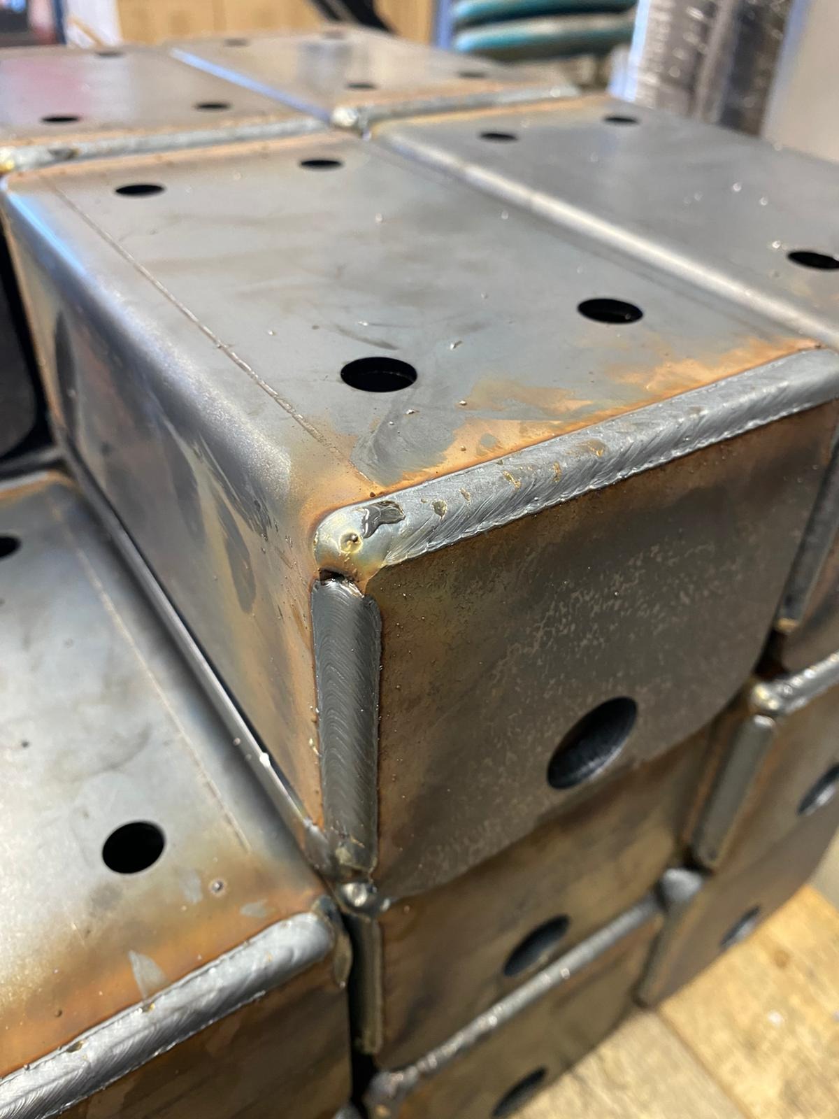 Stos metalowych, spawanych profili zamkniętych z otworami, widoczne ślady spawania i niewielka korozja powierzchniowa.