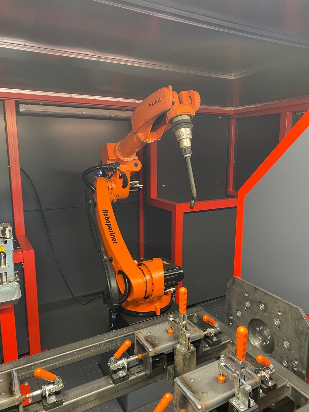 Pomarańczowy robot spawalniczy KUKA Robopartner w kabinie, precyzyjnie ustawiony nad metalowym stanowiskiem z zaciskami, gotowy do automatycznego spawania elementów.
