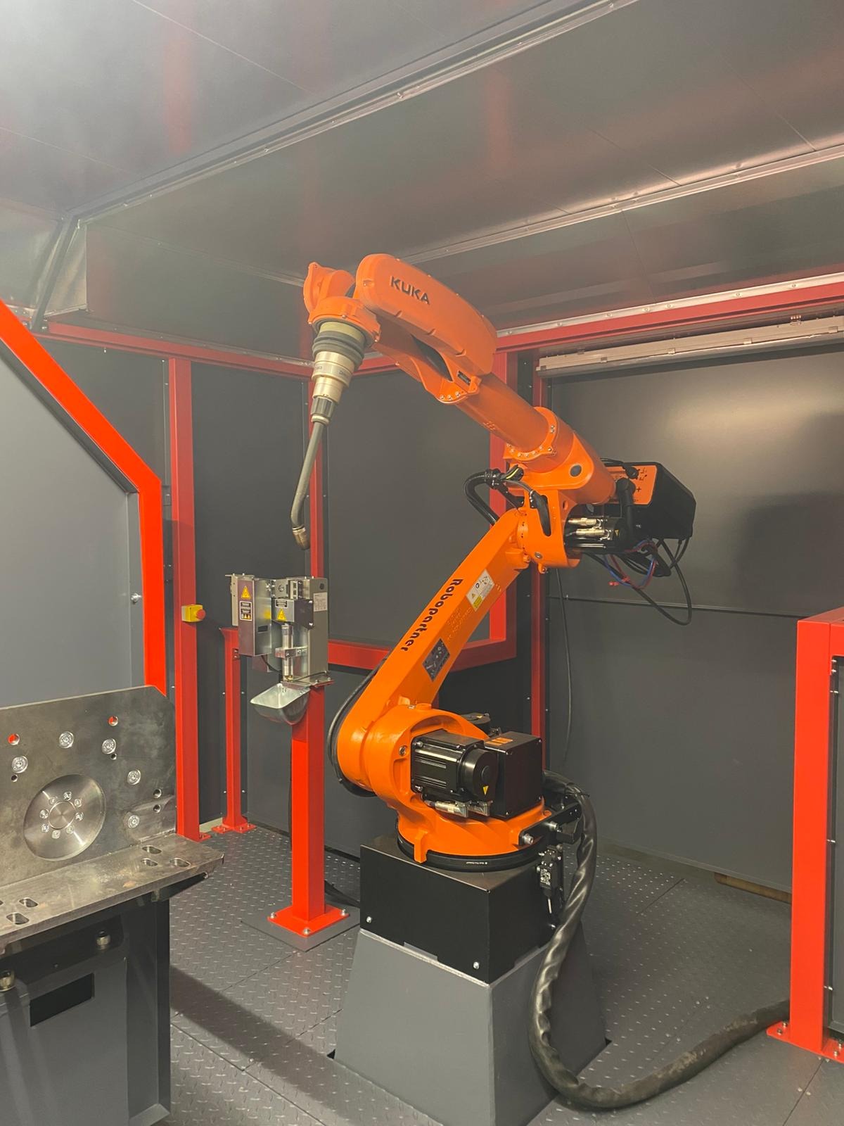 Pomarańczowy robot przemysłowy KUKA w szarej kabinie, z uchwytem narzędziowym skierowanym w stronę stanowiska pracy z metalowymi elementami, czerwona rama konstrukcyjna, stanowisko do obróbki metalu.