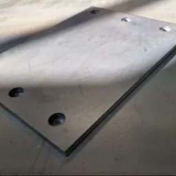 Metalowa płyta z otworami, leżąca na metalowym stole, widoczne ślady obróbki i opalizujące przebarwienia.