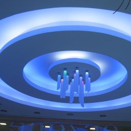 Keras Unibud - Podwieszany sufit z okrągłymi wgłębieniami i niebieskim oświetleniem LED, centralnie umieszczony dekoracyjny żyrandol z podświetlonymi elementami w kolorach niebieskim, zielonym i różowym.