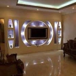 Keras Unibud - Nowoczesna ściana telewizyjna z podświetlanymi półkami gipsowymi w kształcie geometrycznym, błyszcząca podłoga odbija światło, widoczne fragmenty mebli w tle.