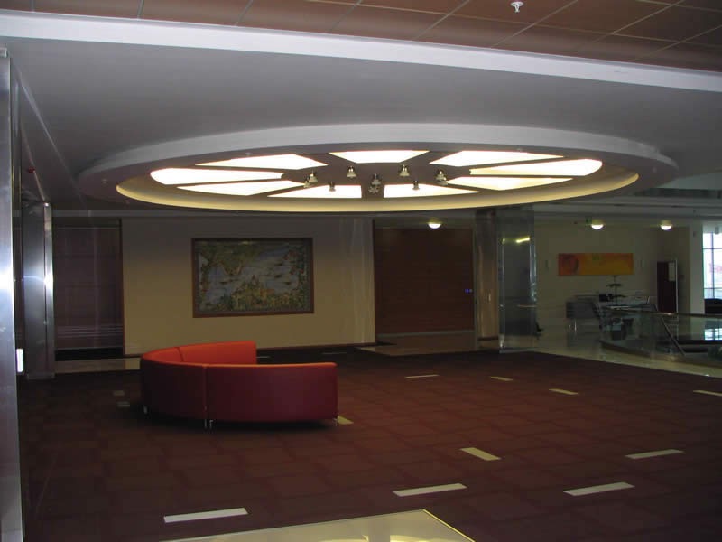 Przestronne wnętrze holu w Pruszkowie z ciemną podłogą, czerwonym półokrągłym siedziskiem i dekoracyjnym, podwieszanym sufitem z oświetleniem.