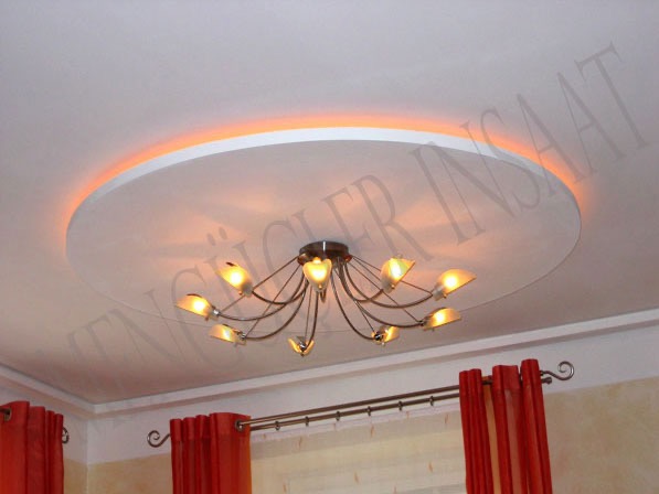 Okrągła podwieszana płyta gipsowa na suficie z oświetleniem LED i ozdobnym żyrandolem, widoczne fragmenty czerwonych zasłon.