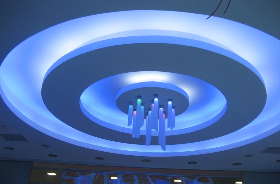 Podwieszany sufit z okrągłymi wgłębieniami i niebieskim oświetleniem LED, centralnie umieszczony dekoracyjny żyrandol z podświetlonymi elementami w kolorach niebieskim, zielonym i różowym.