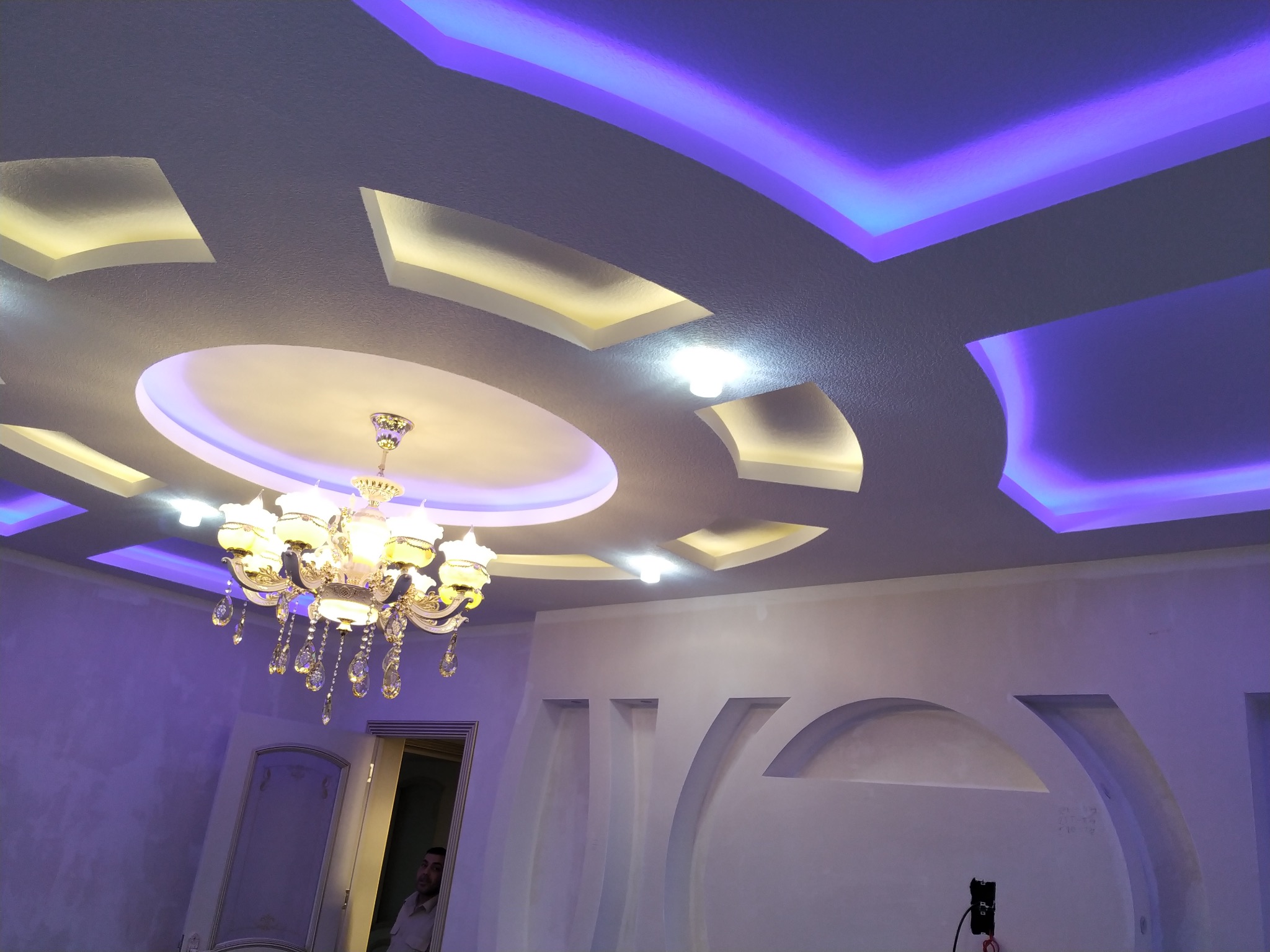 Dekoracyjny sufit podwieszany z oświetleniem LED w kolorze fioletowym i żółtym, oraz kryształowy żyrandol w wykończonym wnętrzu.