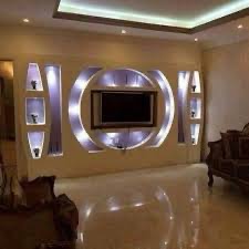 Nowoczesna ściana telewizyjna z podświetlanymi półkami gipsowymi w kształcie geometrycznym, błyszcząca podłoga odbija światło, widoczne fragmenty mebli w tle.