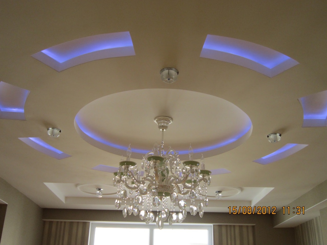 Sufit podwieszany z dekoracyjnym oświetleniem LED w kształcie łuków i okręgu, kryształowy żyrandol w centrum, kremowa kolorystyka.