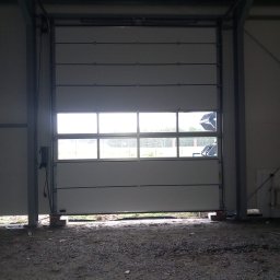 BRAM HOME PIOTR OLEJNICZAK - Biała brama przemysłowa segmentowa z oknami, częściowo podniesiona, widok z wnętrza hali na zewnątrz, w tle widoczny teren z zieleń i sprzęt budowlany.