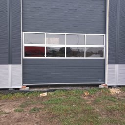 BRAM HOME PIOTR OLEJNICZAK - Szara segmentowa brama przemysłowa z rzędem okien, widoczna z zewnątrz budynku, na nierównym, błotnistym podłożu z fragmentami trawy.