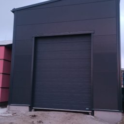 BRAM HOME PIOTR OLEJNICZAK - Zamknięta, segmentowa, antracytowa brama wjazdowa do hali przemysłowej, z widocznymi śladami błota na posadzce przed bramą.