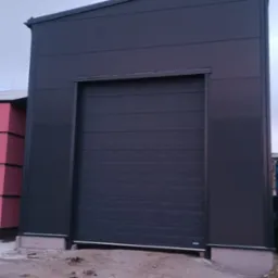 Zamknięta, segmentowa, antracytowa brama wjazdowa do hali przemysłowej, z widocznymi śladami błota na posadzce przed bramą.