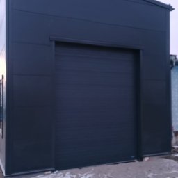 BRAM HOME PIOTR OLEJNICZAK - Zewnętrzny widok nowej, antracytowej bramy segmentowej w budynku przemysłowym, z widocznymi panelami i prowadnicami, na tle zimowego krajobrazu z resztkami śniegu.