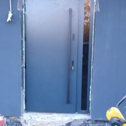 BRAM HOME PIOTR OLEJNICZAK - Szare drzwi zewnętrzne z długim uchwytem i wąskim przeszkleniem bocznym, zamontowane w surowym otworze ściennym z widocznymi śladami pianki montażowej i niedokończonym wykończeniem podłoża.