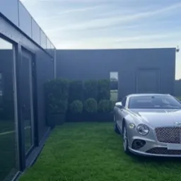 Nowoczesny dom z ciemną elewacją, dużymi oknami i drzwiami, otoczony trawnikiem z r&oacute;wno przyciętymi krzewami, z zaparkowanym srebrnym samochodem Bentley na pierwszym planie.