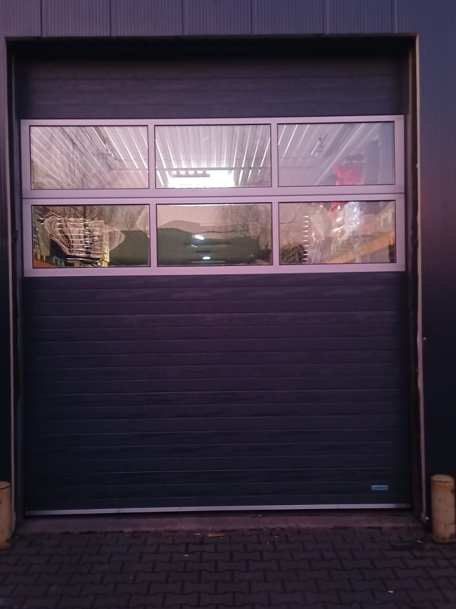 Szara brama segmentowa z sześcioma prostokątnymi oknami w górnej części, widok z zewnątrz na ceglany chodnik.