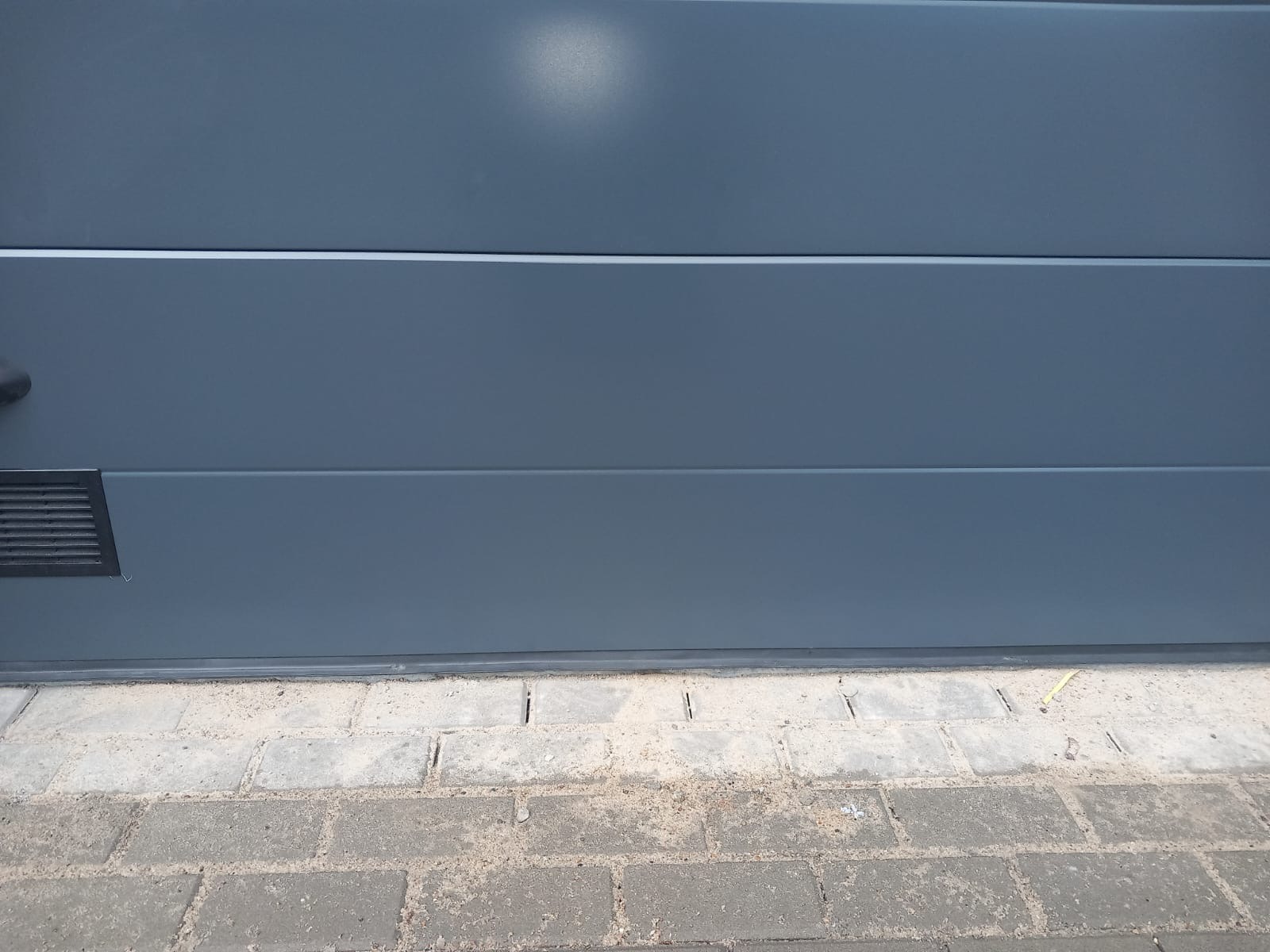 Fragment zamkniętej, szarej bramy segmentowej garażowej z widoczną kratką wentylacyjną w dolnym lewym rogu, uszczelką dolną i kostką brukową przed nią, z leżącym na niej żółtym ołówkiem.