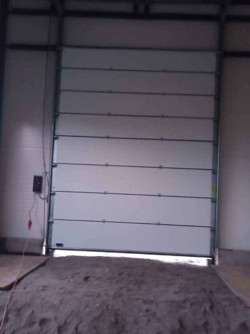 Otwarta, segmentowa brama garażowa wewnątrz hali, widoczny sterownik na ścianie i piasek na posadzce.