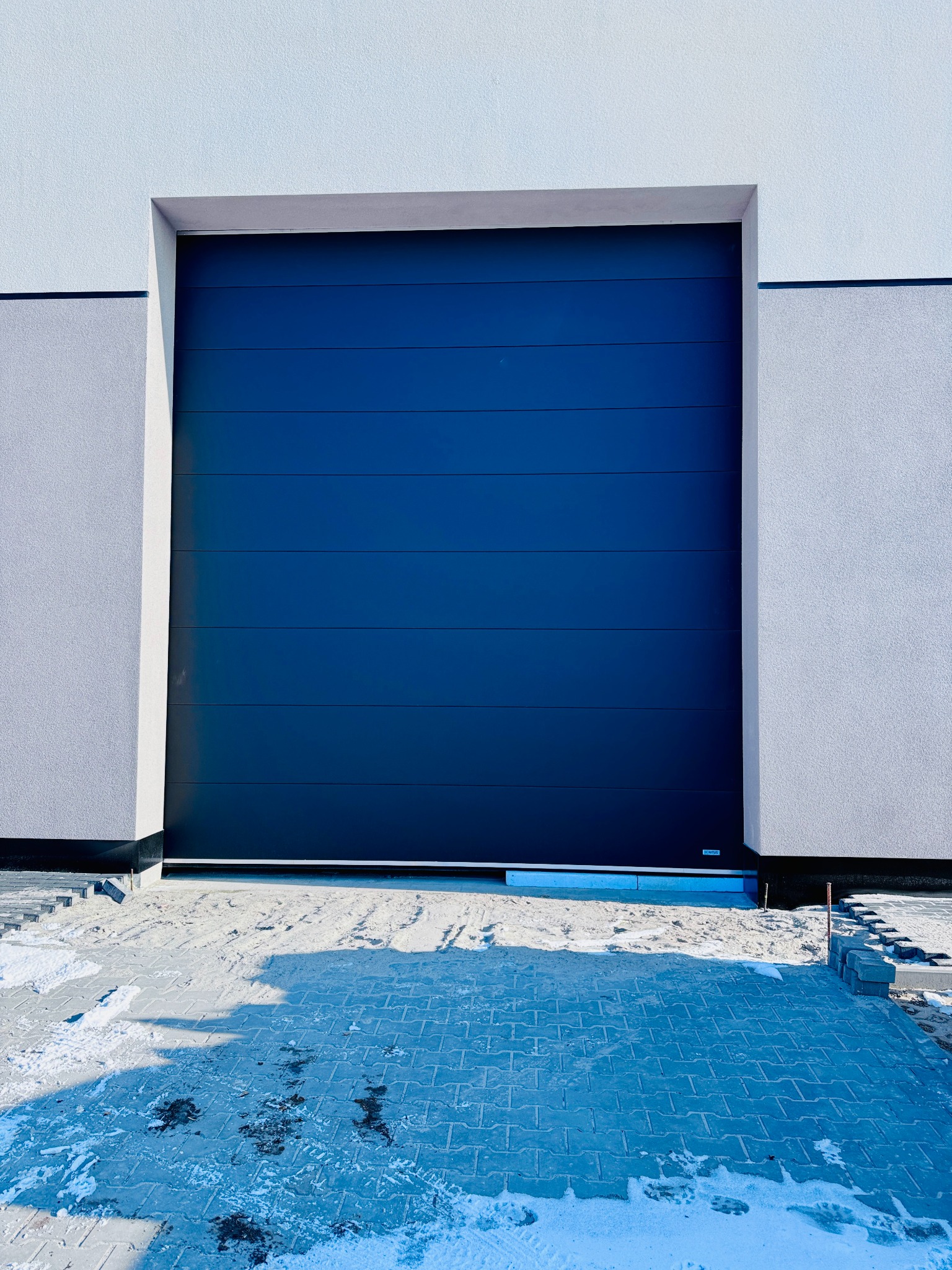 Ciemnogranatowa brama garażowa segmentowa w nowoczesnym domu z jasną elewacją, widoczne ślady śniegu na kostce brukowej przed bramą, brama zamknięta.