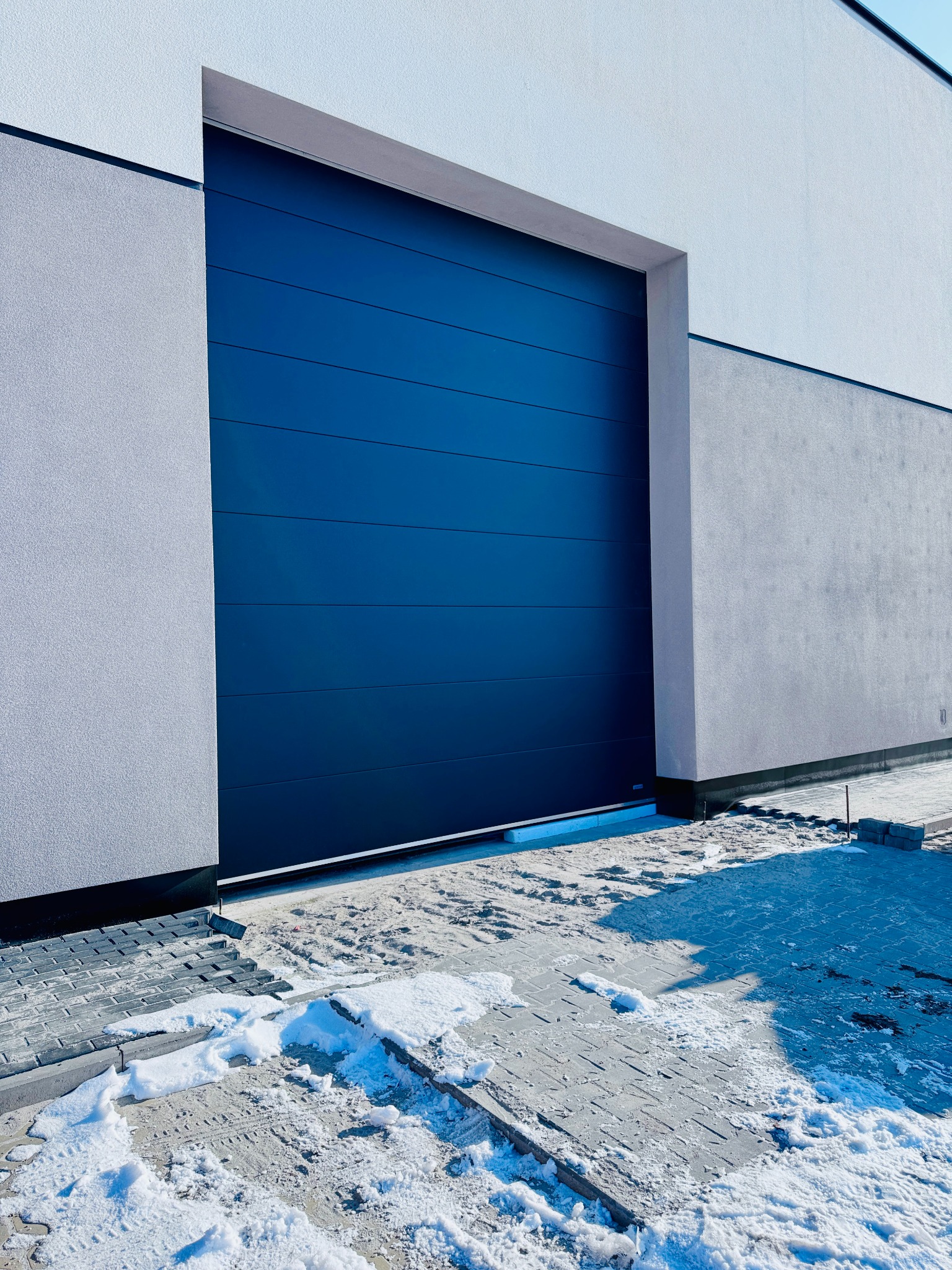 Nowoczesna, segmentowa, ciemnoniebieska brama garażowa, zamontowana w jasnoszarym budynku, z fragmentami śniegu na kostce brukowej przed wjazdem.