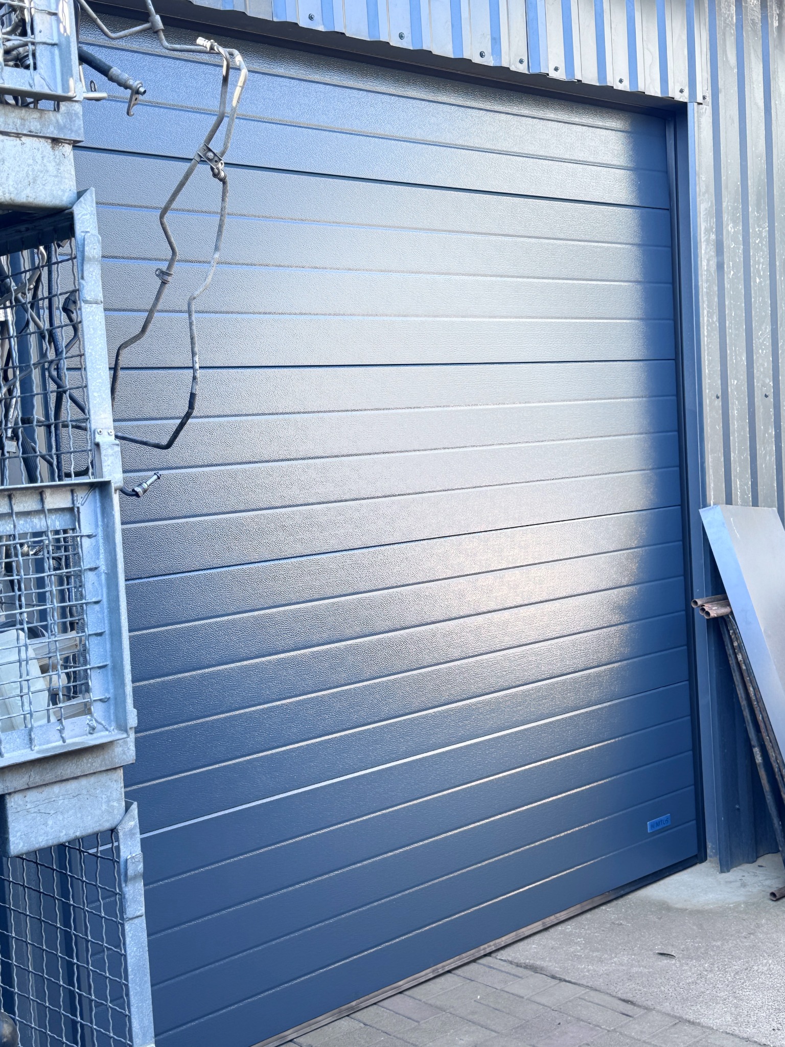 Zamknięta, segmentowa brama garażowa w kolorze grafitowym, z widocznym metalowym koszem i elementami konstrukcyjnymi budynku w tle.