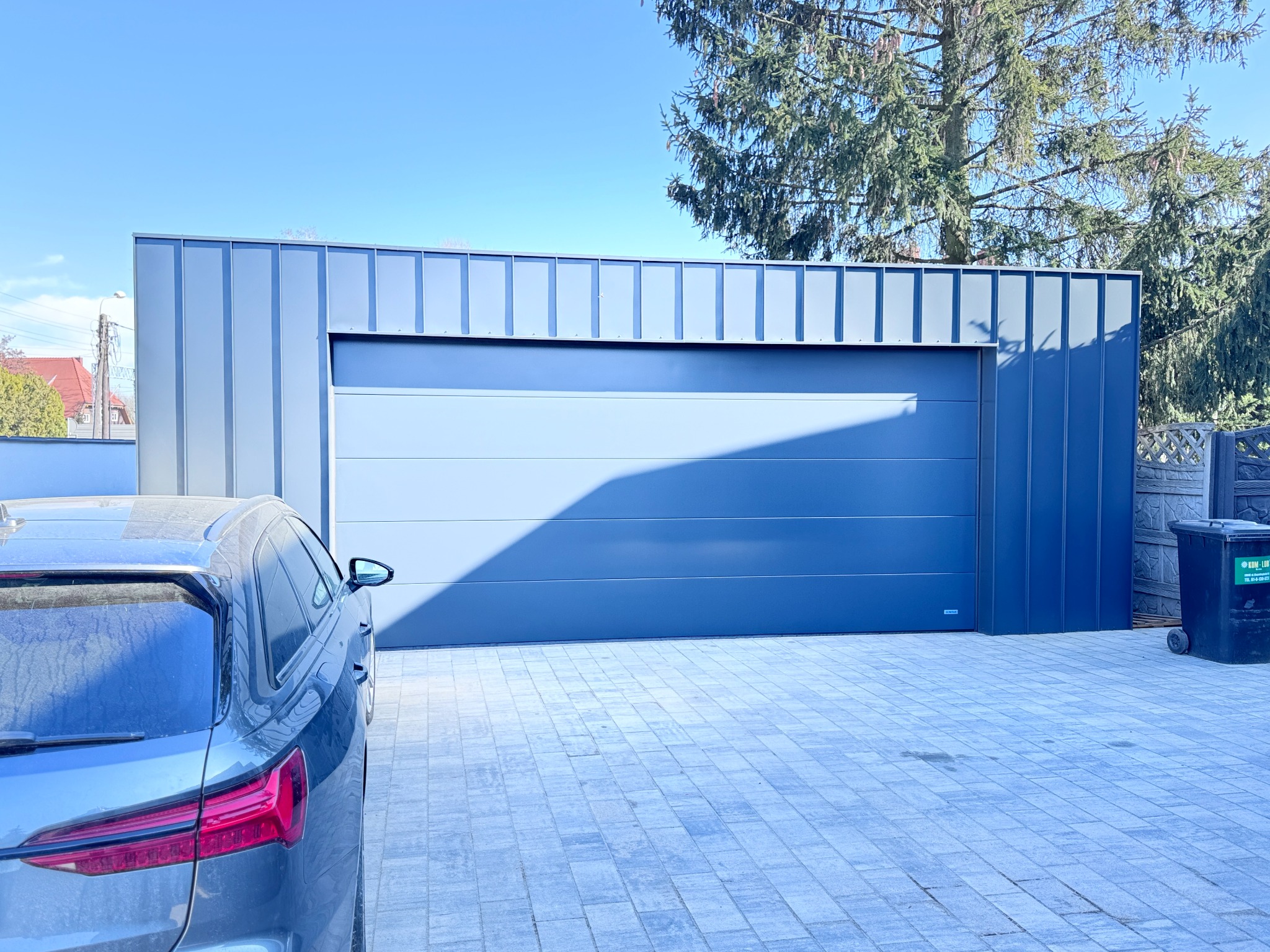 Nowoczesna, segmentowa brama garażowa w kolorze szarym, z widocznym fragmentem samochodu osobowego i kostką brukową na podjeździe, w tle drzewa iglaste i błękitne niebo.