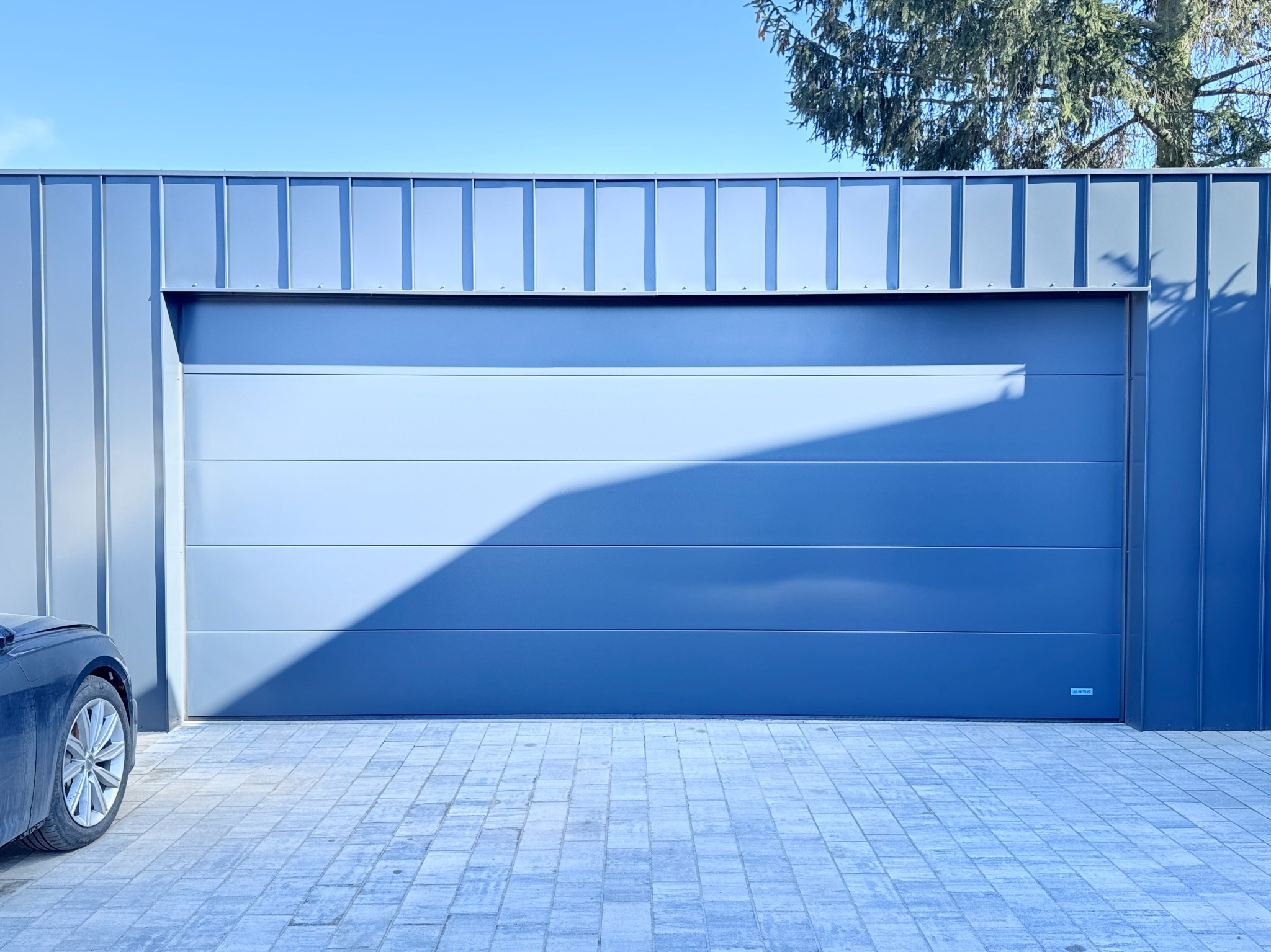 Szara segmentowa brama garażowa, z widoczną częścią zaparkowanego samochodu po lewej stronie, na tle szarej elewacji i kostki brukowej, w słoneczny dzień.