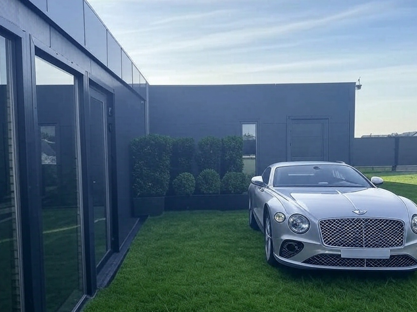 Nowoczesny dom z ciemną elewacją, dużymi oknami i drzwiami, otoczony trawnikiem z równo przyciętymi krzewami, z zaparkowanym srebrnym samochodem Bentley na pierwszym planie.