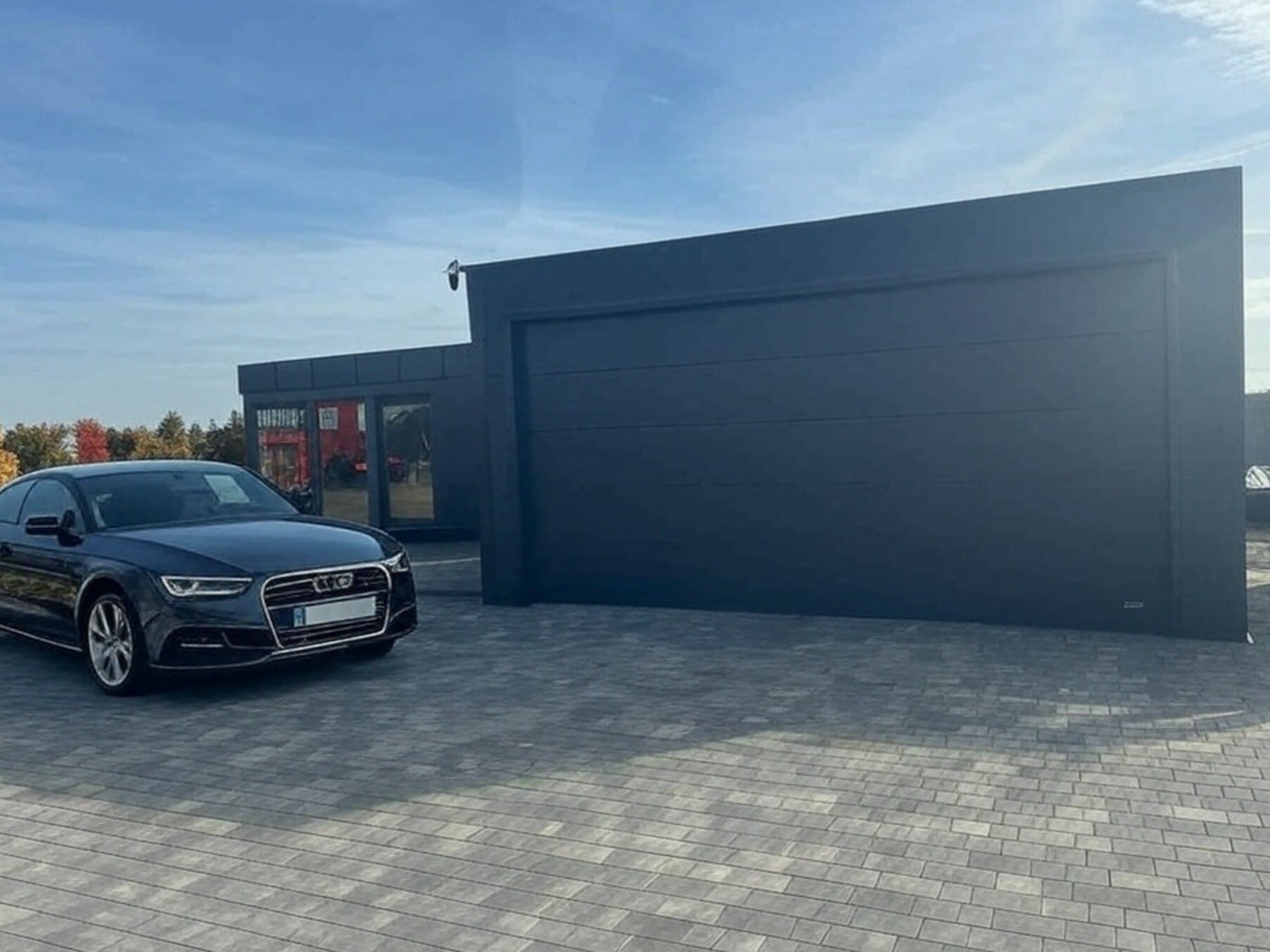 Nowoczesny, antracytowy garaż z bramą segmentową i przyległym budynkiem biurowym, na posesji z szarej kostki brukowej, z zaparkowanym czarnym Audi A4 na pierwszym planie, w słoneczny dzień.